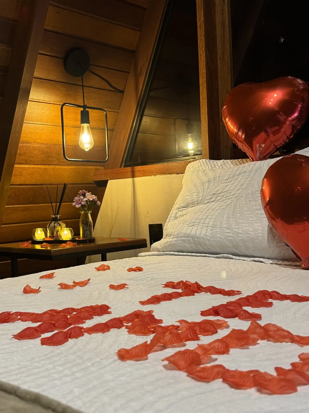 Quarto A-frame com dois balões metalizados de coração, TE AMO em pétalas sobre a cama e velas de LED acesas na cabeceira