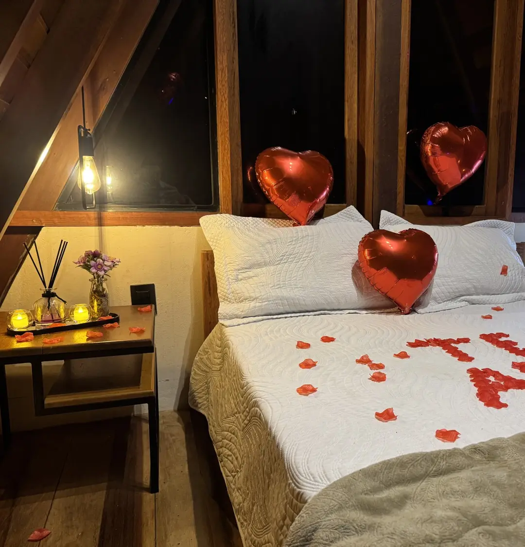 Cama de cabana A-frame com TE AMO escrito em pétalas vermelhas e balões metalizados de coração apoiados nos travesseiros