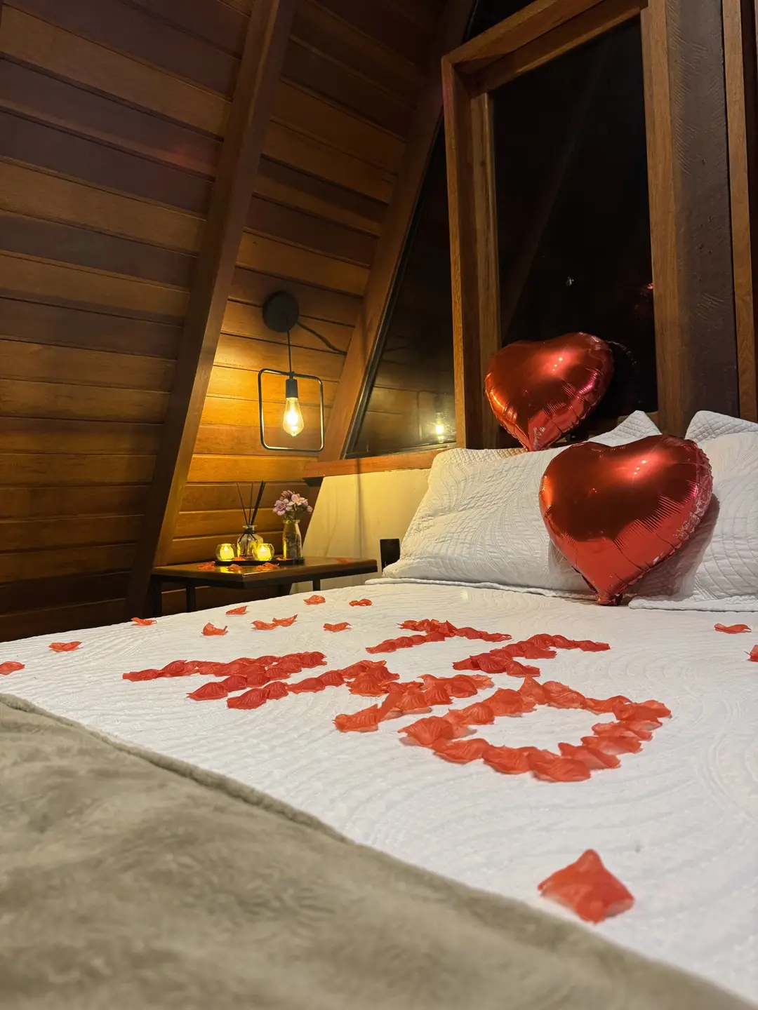 Cama arrumada com TE AMO escrito em pétalas vermelhas e um balão metalizado de coração apoiado no travesseiro