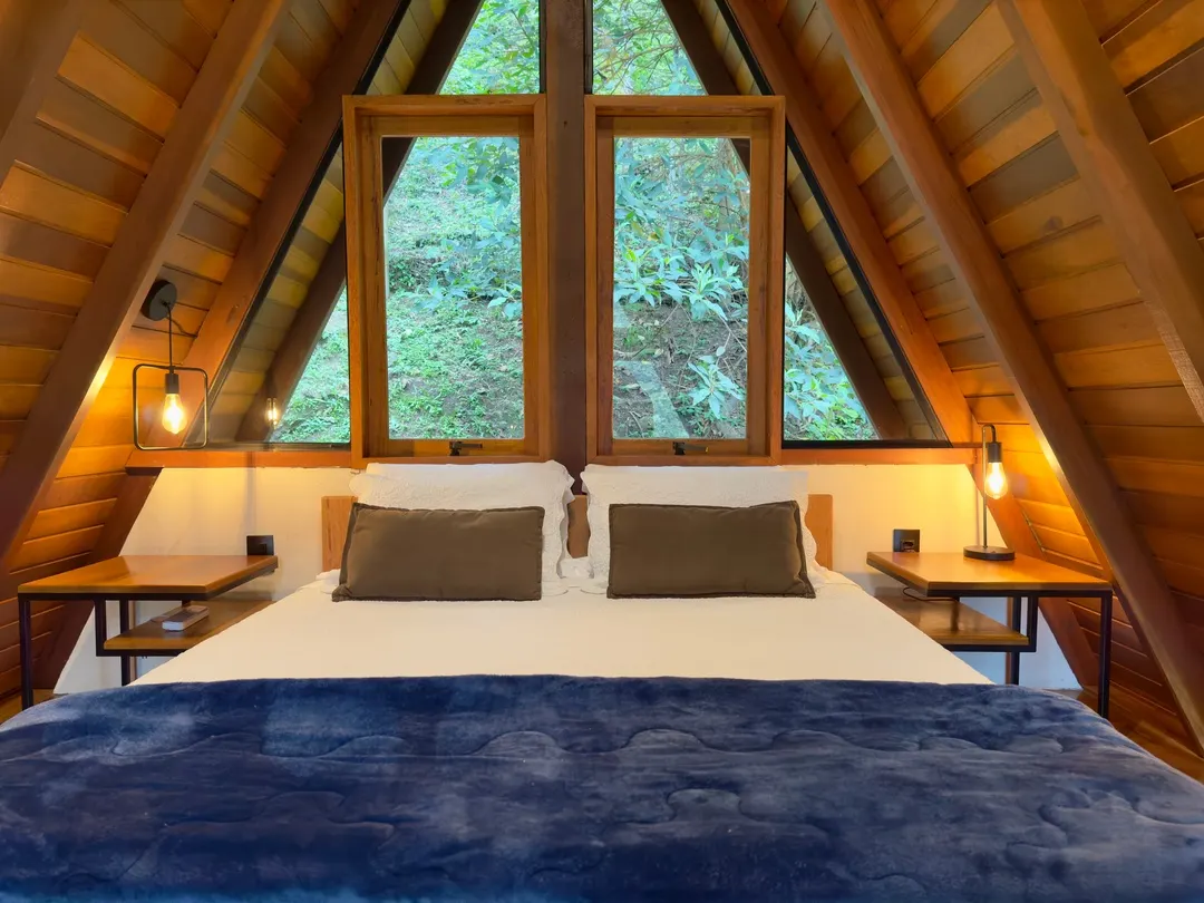 Cama queen da Cabana Sirius entre duas janelas A-frame emolduradas pela mata, luz da manhã