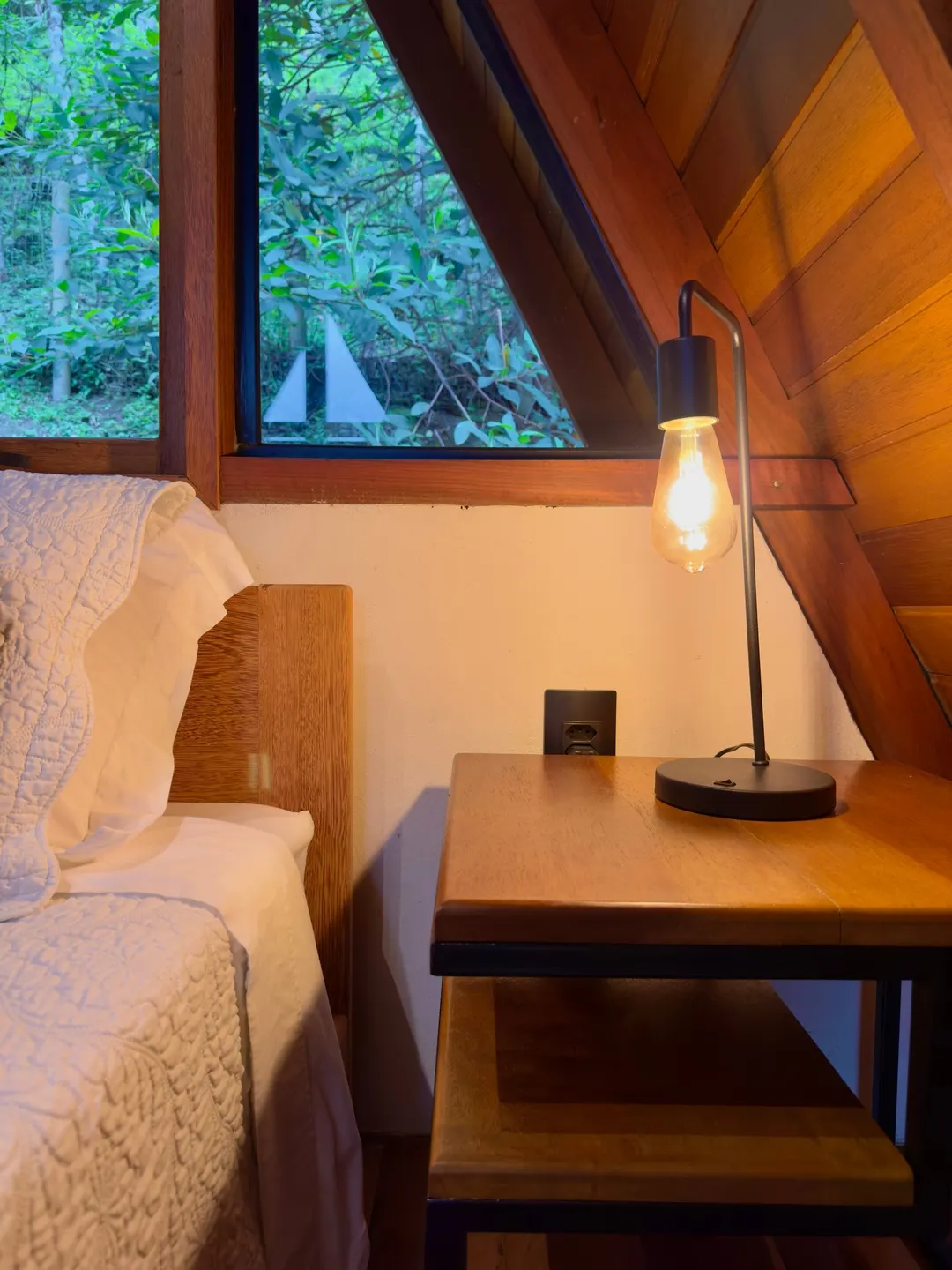 Cama arrumada no mezanino do A-frame, com a mata verde nas janelas triangulares
